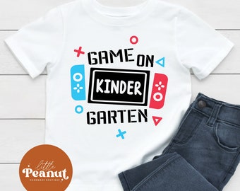 Può includere: Maglietta bianca con una grafica di un controller per videogiochi con il testo "Game On Kinder Garten".