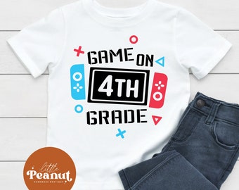 Può includere: Maglietta bianca con un design grafico di un controller per videogiochi con il testo "Game On 4th Grade".