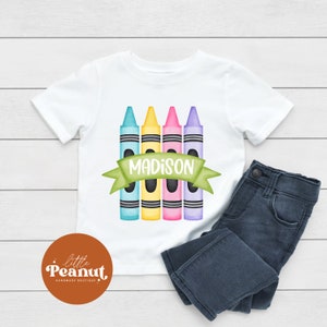 May include: White t-shirt with a design of four colorful crayons and a green banner with the name "MADISON" on it. The t-shirt is laying on a white wooden surface with a pair of blue jeans next to it.