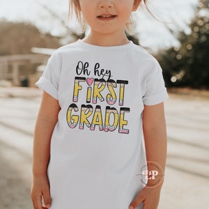 May include: White t-shirt with the text "Oh hey FIRST GRADE" in black and yellow with a pink heart. The letters are made to look like pencils.