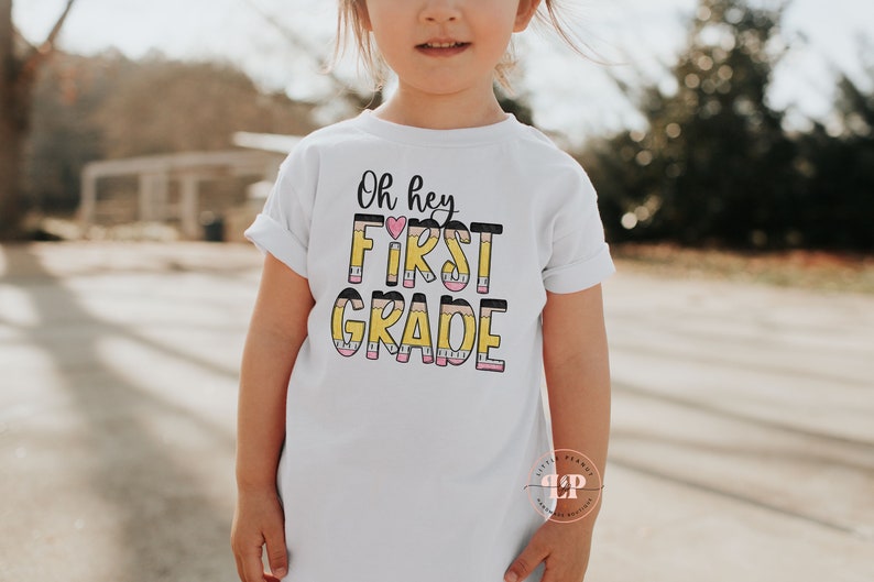 May include: White t-shirt with the text "Oh hey FIRST GRADE" in black and yellow with a pink heart. The letters are made to look like pencils.