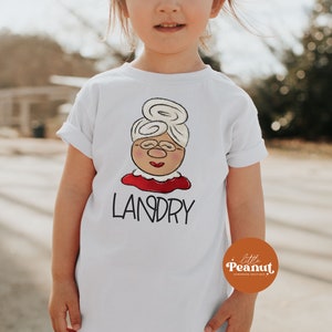 May include: White t-shirt featuring a cartoon illustration of a woman with glasses, a bun hairstyle, and a red collar. The name "LANDRY" is printed below the illustration in black letters. The shirt is for children.