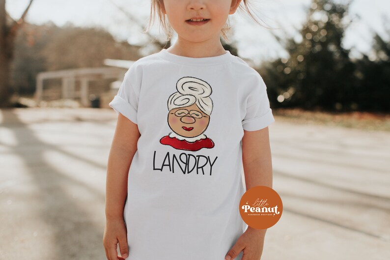 May include: White t-shirt featuring a cartoon illustration of a woman with glasses, a bun hairstyle, and a red collar. The name "LANDRY" is printed below the illustration in black letters. The shirt is for children.