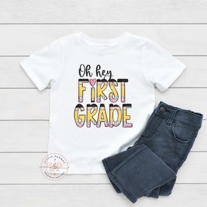 May include: White t-shirt with the text "Oh hey FIRST GRADE" in black and yellow with a pink heart. The text is stylized to look like pencils.