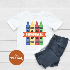May include: White t-shirt with a design of four colorful crayons and the name "Maverick" in a red banner.