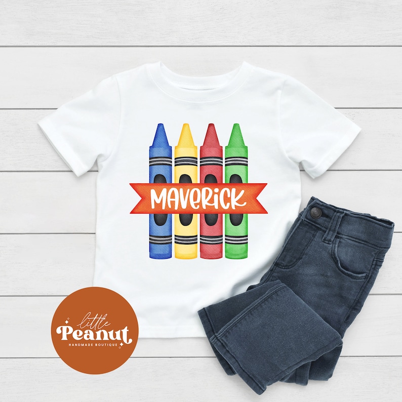 May include: White t-shirt with a design of four colorful crayons and the name "Maverick" in a red banner.