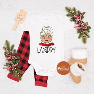 May include: A white baby onesie with a cartoon Mrs. Claus face and the name "LANDRY" in black. Red and black plaid pants and tan baby booties with white fur trim are also in the image. A wooden rattle and holiday greenery complete the festive scene.
