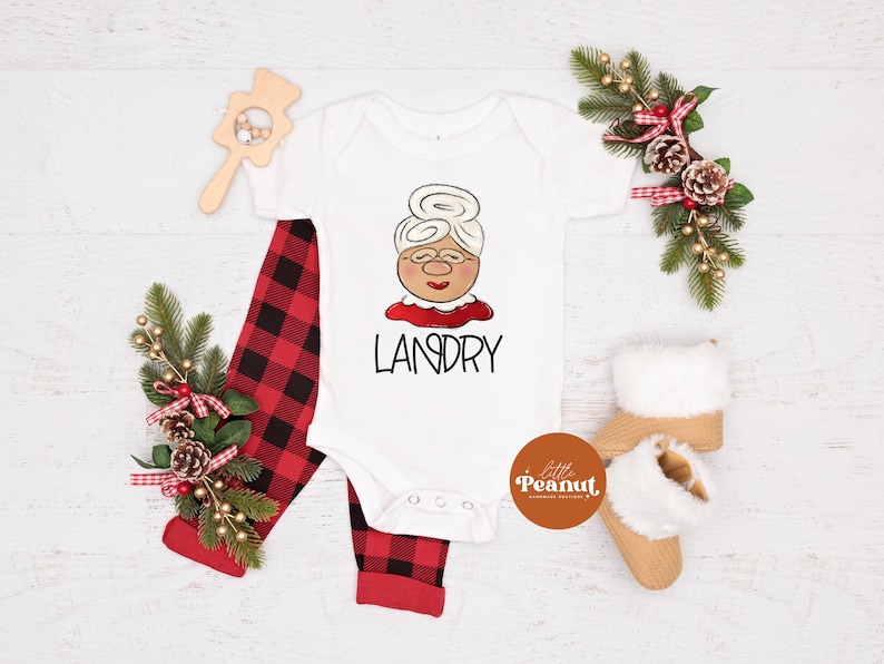 May include: A white baby onesie with a cartoon Mrs. Claus face and the name "LANDRY" in black. Red and black plaid pants and tan baby booties with white fur trim are also in the image. A wooden rattle and holiday greenery complete the festive scene.
