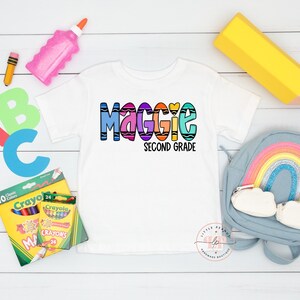 May include: White t-shirt with a colorful crayon-themed design that says "Maggie Second Grade". The shirt is surrounded by school supplies, including crayons, a pencil, a glue stick, and a backpack.