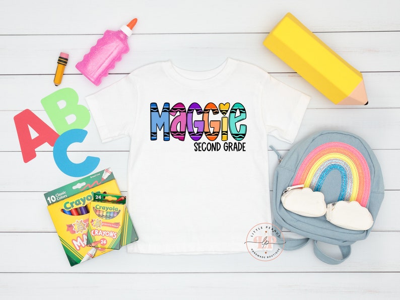 May include: White t-shirt with a colorful crayon-themed design that says "Maggie Second Grade". The shirt is surrounded by school supplies, including crayons, a pencil, a glue stick, and a backpack.