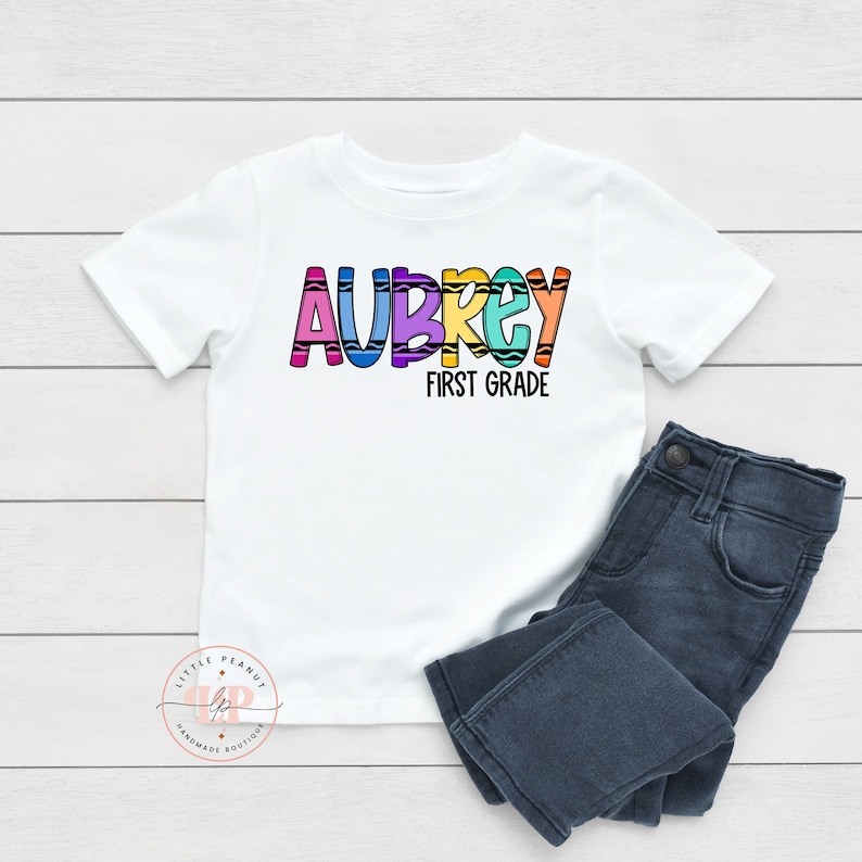 May include: White t-shirt with the name "Aubrey" spelled out in colorful crayon-like letters. The text "First Grade" is below the name.