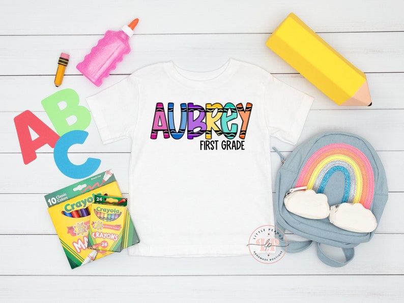 May include: White t-shirt with the name "Aubrey" in rainbow crayon-like letters and the text "First Grade" below. The shirt is surrounded by school supplies, including crayons, a pencil, a glue stick, and a backpack.