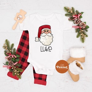 May include: White baby onesie with a cartoon Santa Claus face and the name "Luke" printed on it. The onesie is paired with red and black plaid pants.