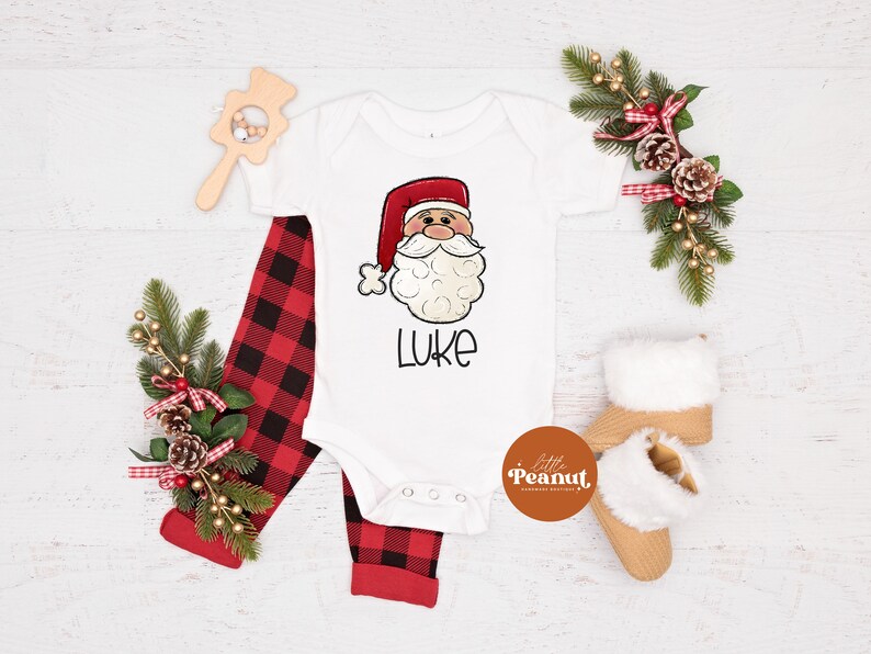 May include: White baby onesie with a cartoon Santa Claus face and the name "Luke" printed on it. The onesie is paired with red and black plaid pants.