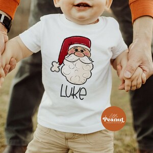 May include: A white t-shirt with a cartoon Santa Claus face and the name "LUKE" printed on it. The shirt is being held by a child and an adult.