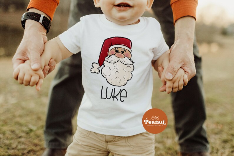 May include: A white t-shirt with a cartoon Santa Claus face and the name "LUKE" printed on it. The shirt is being held by a child and an adult.