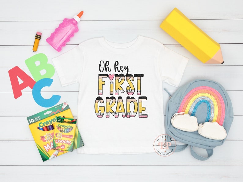 May include: A white t-shirt with the text "Oh hey FIRST GRADE" in black and pink letters with a yellow pencil design. The shirt is surrounded by school supplies, including crayons, a pencil, a glue stick, a backpack, and a large yellow pencil.