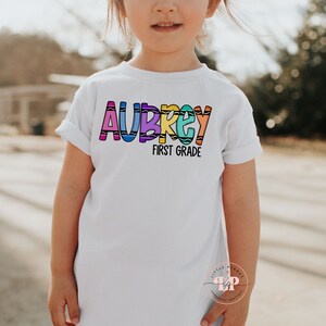May include: A white t-shirt with the name "Aubrey" spelled out in colorful crayon-like letters. The text "First Grade" is written below the name.