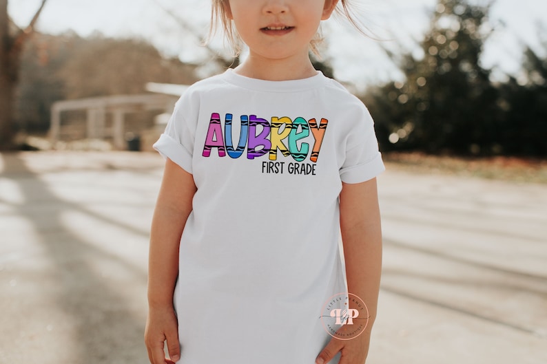 May include: A white t-shirt with the name "Aubrey" spelled out in colorful crayon-like letters. The text "First Grade" is written below the name.