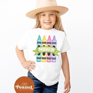 May include: White t-shirt with a design of five colorful crayons and a green banner with the name "MADISON" on it. The shirt is worn by a child wearing a brown hat and blue jeans.