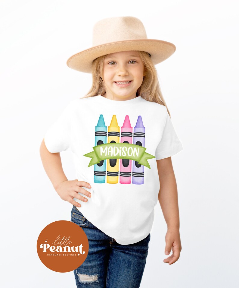 May include: White t-shirt with a design of five colorful crayons and a green banner with the name "MADISON" on it. The shirt is worn by a child wearing a brown hat and blue jeans.