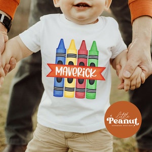 May include: A white t-shirt with a design of four colorful crayons and a red banner with the name "Maverick" printed on it. The shirt is being held by two hands.