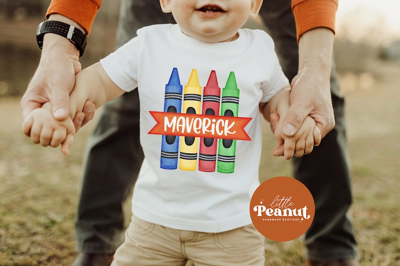 May include: A white t-shirt with a design of four colorful crayons and a red banner with the name "Maverick" printed on it. The shirt is being held by two hands.