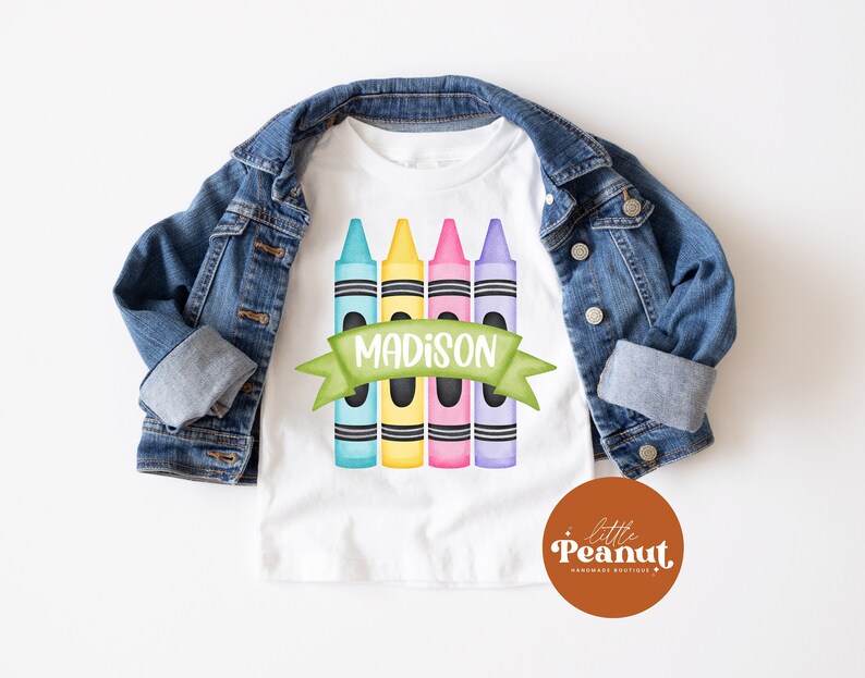 May include: White t-shirt with a design of five colorful crayons and a green banner with the name "MADISON" on it.