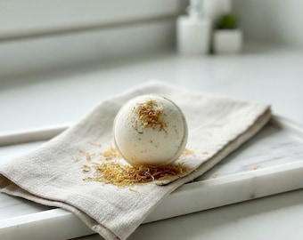 May include: A white bath bomb with flecks of dried petals sits on a folded linen cloth. The cloth rests on a white marble tray, with scattered petals around the bath bomb. The scene is set against a bright, neutral background.