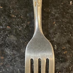 Può includere: Una forchetta in metallo color argento con una figura in rilievo di un personaggio con mantello sulla parte superiore del manico. La parola BATMAN è incisa verticalmente in lettere maiuscole sotto la figura, lungo il centro del manico.