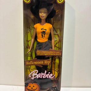 May include: A Barbie doll in original packaging, featuring a black hat, an orange t-shirt with a black cat graphic, and a long black skirt. The box displays the text Halloween Hip, Barbie, and What a treat! Bracelets for you!, with a pumpkin illustration.