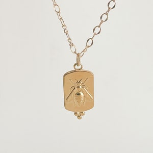 May include: Gold necklace with a rectangular pendant featuring a detailed bee design. The pendant has three small beads hanging from the bottom.