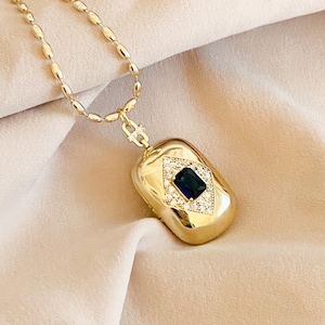 May include: A gold-toned necklace with a rectangular pendant. The pendant features a dark blue gemstone in a diamond shape, surrounded by smaller clear stones. The chain has an oval link design. The jewelry is displayed on a beige fabric.