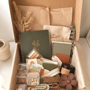 May include: A cardboard gift box containing a dark green spiral notebook with floral gold foil, a smaller green notebook, a black candle labeled Caramel Apple, a brown flower hair clip, clear plastic containers with stationery, and paper items.