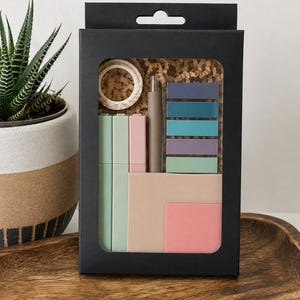 May include: A black rectangular box containing a stationery set with a roll of patterned washi tape, a neutral-colored pen, three pastel-colored markers, a stack of six rectangular sticky note tabs, and two square sticky note pads in beige and pink.