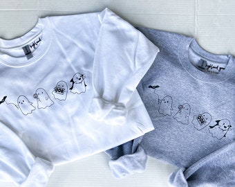 May include: Two sweatshirts, one white and one gray, featuring a line of cartoon ghosts with Halloween-themed designs. The ghosts are holding a bat, a lollipop, a sign that says "BOO", and a vampire cape. The text "Highland Grove Designs" is printed on the top right.