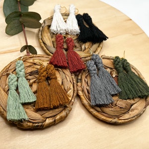 May include: Assortment of handmade tassel earrings in various colors, including green, brown, red, gray, black, and white. The earrings are displayed on woven mats.