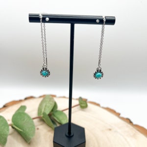 May include: A pair of silver chain earrings with turquoise stones set in a silver bezel. The earrings are hanging from a black stand.