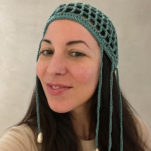 Peut inclure: Un couvre-chef en crochet bleu sarcelle avec un motif en maille ouverte sur le dessus. L'accessoire comprend quatre longs liens tressés pendants qui descendent sur les côtés, chacun terminé par un coquillage blanc à l'extrémité inférieure.