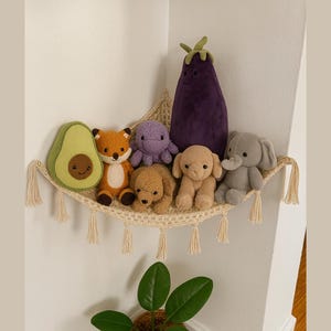 Puede incluir: Una estantería de macramé beige exhibe peluches, incluyendo un aguacate, un zorro, un pulpo, perros, un elefante y una berenjena. Los juguetes son de varios colores, como verde, naranja, morado, marrón y gris. La estantería está decorada con borlas y está montada en una pared blanca.