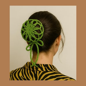 Pode incluir: Acessório de cabelo verde de crochê, com um design circular vazado e um cordão verde a condizer amarrado em laço. A pessoa está vestindo uma camisa listrada em preto e amarelo. O cabelo é escuro.