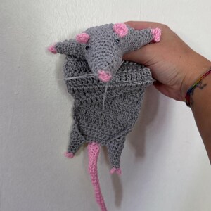 May include: A handmade crochet rat toy in gray with pink accents. The rat has pink ears, feet, and a long pink tail. The toy is held against a white wall.