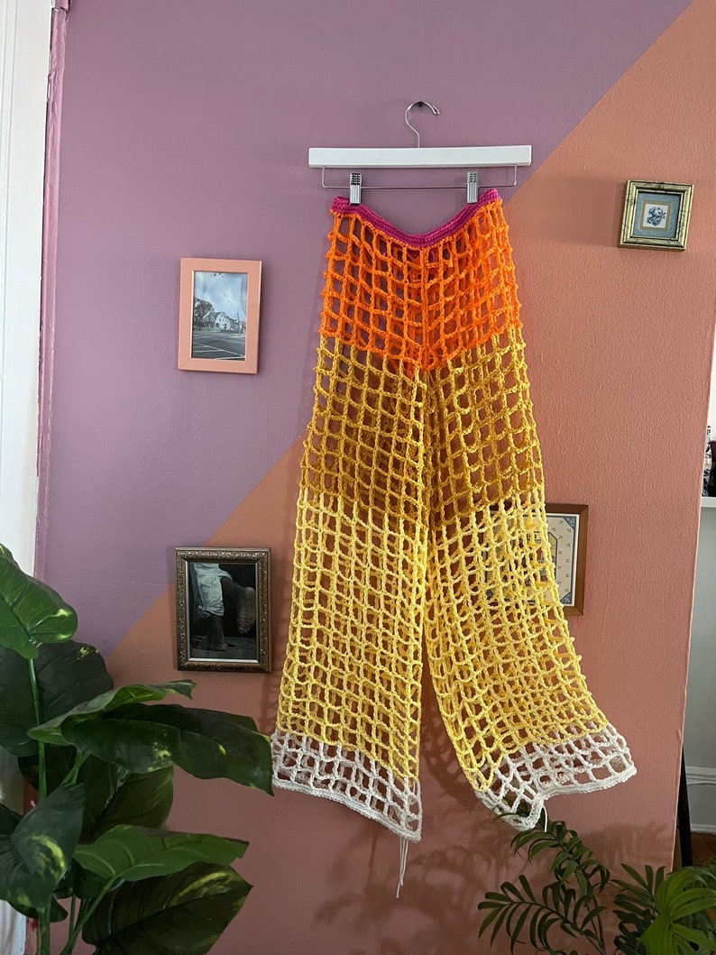 May include: Crocheted pants with a wide leg, featuring a multi-colored design. The pants are made of a lightweight, airy material and have a relaxed fit. The colors are pink, orange, yellow, and white.