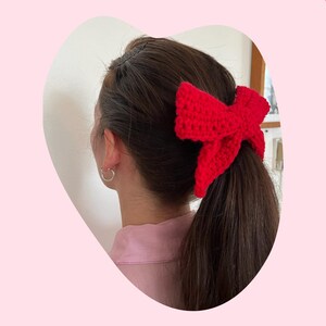 May include: A red crocheted bow hair tie. The bow is tied around a ponytail.