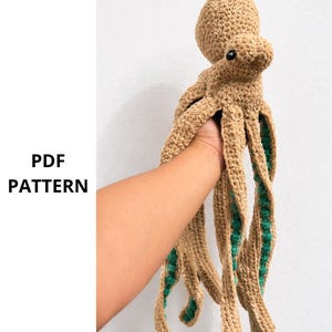 May include: A crocheted octopus in shades of tan and green. The octopus has a rounded head, a body, and eight long tentacles. The tentacles are tan with green accents. The text "PDF PATTERN" is on the left side of the image.