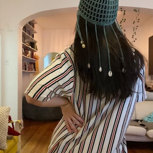 May include: A teal crocheted hat with a net-like design and dangling strands adorned with small white shells. The hat is worn over long, dark hair. The person is wearing a striped dress with red and black accents.