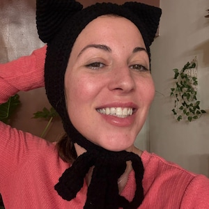 May include: A black crochet cat ear hat with a tie closure. The hat features two upright ears and a tie that hangs down. The hat is made of a soft, textured yarn. The hat is worn by a person wearing a pink shirt.