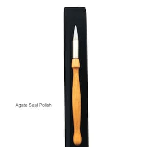 May include: A wooden handled agate seal polish applicator in a black box. The applicator has a white, pointed tip. The text "Agate Seal Polish" is visible below the applicator. The text "Calligraphy Story" is visible below the box.