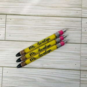 May include: Three yellow pencil-shaped pens with black glitter and pink accents. Each pen has the text "Ms. Avalyn" written in black.