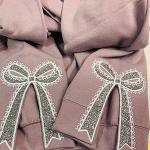 May include: Two mauve sweatshirts with a white and silver glitter bow design on the sleeves.
